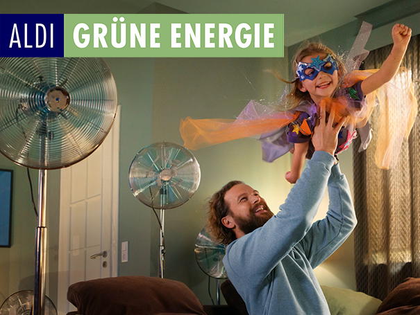 ALDI GRÜNE ENERGIE. Vater hebt Tochter in Superwoman Kostüm in die Luft.